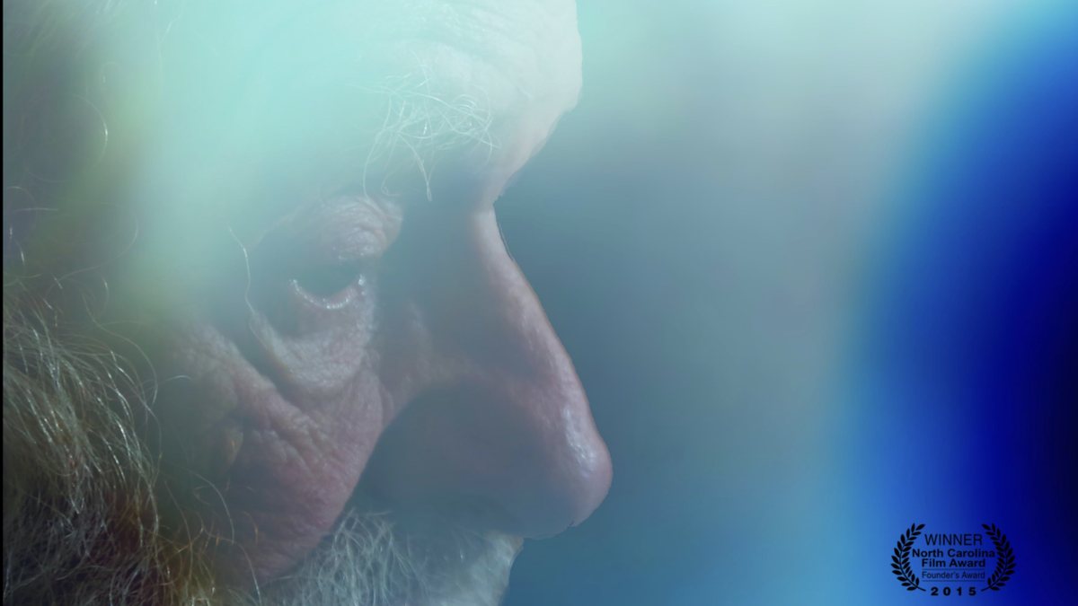 An elderly man in soft-focus profile against a pale blue background, from the In Memory poster