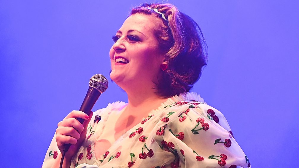 A comedian in a cherry-print dress laughing at the mic under blue stage light