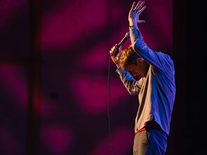 Stand-up performer mid-routine against a magenta backdrop